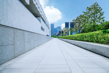 Empty stone footpath alongside a modern building exterior with green landscaping and city skyline.