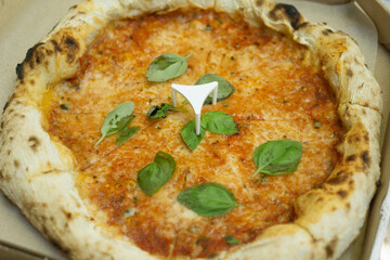 overhead close up of a whole baked margherita pizza with fresh basil and melted cheese in a cardboard takeout box concept of food delivery, pizzeria advertising, casual dining