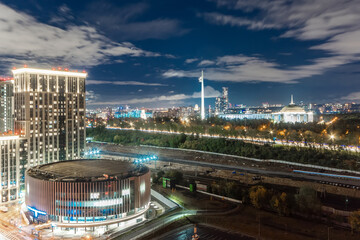 A bustling cityscape showcases dazzling lights from modern buildings and roads at night, with a serene park in the foreground framed by trees and distant architecture