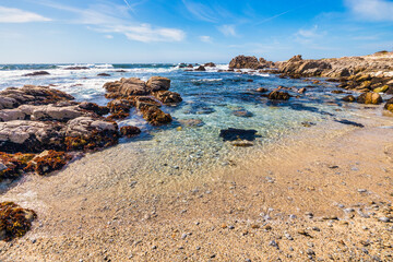 Rocky shoreline along 17 Mile Drive near Monterey, California. Waves crash against rugged stones, with coastal plants adding color under a bright sky on this famous scenic route by the Pacific Ocean