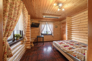 Cozy log cabin bedroom with wooden walls, ceiling, and dark floor. A single bed with a colorful quilt, a small table, a TV, and floral curtains by the window