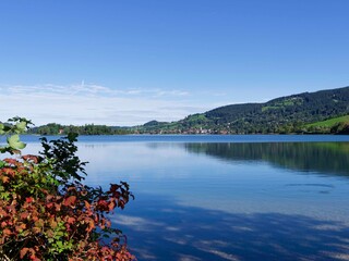 The calm waters of Lake Schliersee under a blue sky facing the town of Schliersee and its bell tower on the northeast horizon between the slopes of the Schliesberg and Huberspitz

