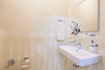 A well-lit bathroom features beige tiled walls, a sleek white sink, and a mirror. Chrome fixtures and a towel add a touch of modern comfort