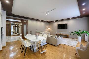 A modern living room with a dining area featuring a white table and chairs. The room has hardwood floors, gray walls, a beige sofa, and a large TV. A mirror closet is present