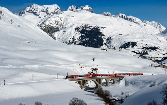 Aerial view of a red train gracefully traversing a snow-laden viaduct, framed by the majestic, snow-covered Alps under a clear sky, Filisur, Switzerland.