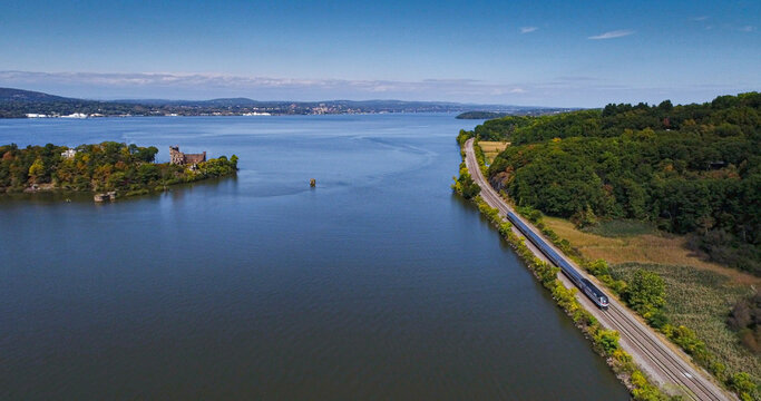 Aerial view of a sleek train glides along the tracks beside a tranquil lake, with a historic island castle standing proudly, Hudson River