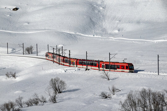 Aerial view of a vibrant red train slicing through a pristine white snowscape, winding past the stark, bare trees, Filisur, Switzerland.