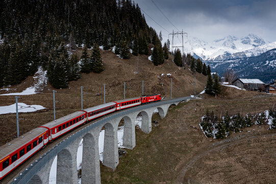 Aerial view of a vibrant red train gracefully traverses the majestic Landwasser Viaduct against a backdrop of snow-dusted mountains, Filisur, Switzerland.