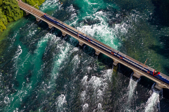 Aerial view of a train traversing a stone bridge over the churning turquoise waters of a rapid river, contrasting with the dark depths below, Schaffausen falls
