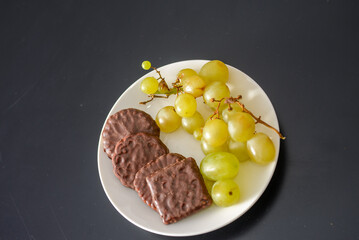 White grapes and chocolate biscuits on the same plate as two contrasts of healthy and unhealthy sugars and so-called treat foods on a dark background