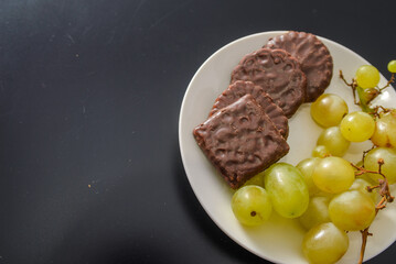 White grapes and chocolate biscuits on the same plate as two contrasts of healthy and unhealthy sugars and so-called treat foods on a dark background