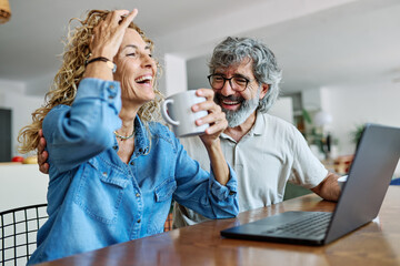 Portrait of a lovely senior mature couple using a laptop together and having fun drinking coffee or tea sitting at a table at home