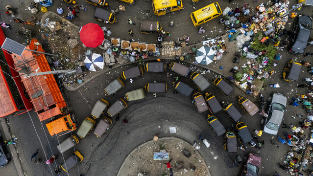 Aerial view of yellow and brown danfo buses and Keke Napeps converge in a bustling roundabout, framed by vibrant market stalls and scattered waste, Lagos, Lagos, Nigeria.