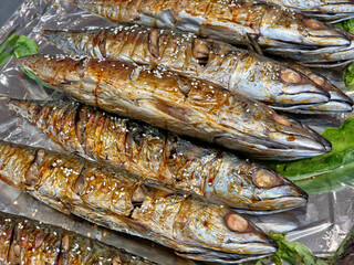 Close-up of several grilled mackerel arranged on foil, scored and glazed with sauce, topped with sesame seeds, and garnished with greens.