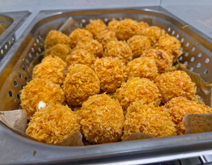 numerous  deep-fried, cheese balls, piled inside a perforated, stainless steel catering tray.