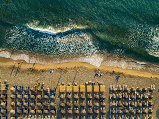 Aerial view of sun-soaked beach umbrellas line the golden sands where foamy waves crash ashore, painting a picturesque scene of seaside bliss, Chania, Chania, Greece.