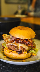 A succulent grilled burger with melted cheese, crispy bacon, sweet corn, and fresh salad on a white plate. Perfect homemade meal, top view.