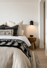 a bedroom with white walls, a beige bed and black patterned bedding, an illuminated sconce on the wall above it, a small wooden stool beside the bedside table