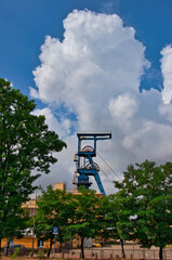 
Wysoka, niebieska wieża g&oacute;rnicza wznosi się nad porośniętym drzewami kompleksem przemysłowym na tle jasnego nieba. 