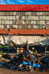 Opuszczona fasada fabryki ukazuje rozbite okna i zardzewiałe panele, a na pierwszym planie dominuje chaotyczna sterta śmieci i gruzu. Scena ukazuje opuszczenie i miejski rozkład pod czystym niebem. © Pol Badger