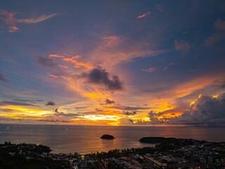 A breathtaking sunset paints the sky in vivid hues of orange and blue over a coastal town, with lush green hills framing the scene. A distant hilltop structure adds a serene tranquil landscape.