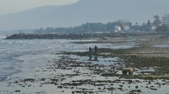 A misty coastal scene during low tide, with people walking across wet sand and scattered rocks. Reflections shimmer in shallow water, while distant breakwater, cars, and houses frame the horizon.