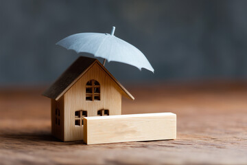 A small wooden house stands on a table, protected by a miniature umbrella, symbolizing safety and security. Wooden block in front.