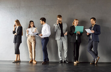 Obraz premium Portrait of confident business people in formal clothes talking on a meeting indoors. Company employees team standing in a row on dark gray wall background communicating with each other.