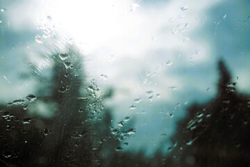 Abstraktes Landschaftsmotiv mit Regentropfenmuster auf Glasscheibe vor hohen grünen Bäumen und blau-grau-weißem Himmel mit Licht in der Natur bei Regen und Sturm am Morgen im Herbst