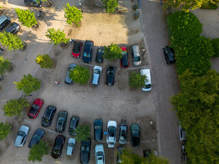 Aerial view of unpaved parking lot with scattered cars, trees, and cobblestone path, showing organized and irregular sections amid surrounding greenery.