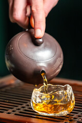 Close-up of hand pouring amber tea from traditional clay teapot into glass cup on wooden tray. Chinese tea ceremony, warm tones, cultural ritual, lifestyle concept