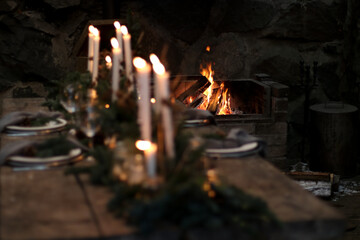 Atmospheric evening scene with stacked firewood and burning fire. A table with candles, beautiful plates and cutlery creates a warm and cozy ambiance.