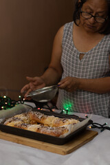 Woman adding powdered sugar to a freshly baked king's cake in a kitchen, celebrating holiday traditions and enjoying a sweet homemade dessert for family gatherings