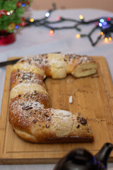 King's cake dusted with powdered sugar and chopped pecans on a wooden board, festive holiday pastry with blurred string lights and a small christmas tree in the background