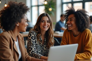 Three diverse women collaborating and laughing together at a modern co-working space, enjoying positive teamwork