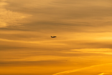 Silhouette of a passenger airplane flying through a vast and beautiful golden orange sky at sunset or sunrise.
