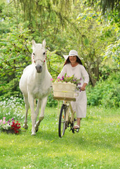 Woman riding a white bicycle in the garden, smiling, horse running near her towards camera