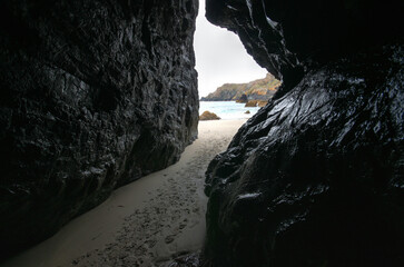 Narrow cave like passage between large dark rocks on smooth sand beach at Kynance Cove, footprints over ground, ocean in background