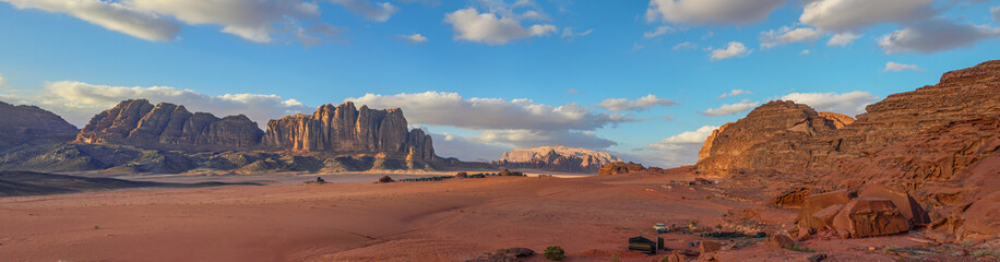 Wadi Rum desert with red sand lit by evening sun, rocky mountain formations in background, camp near