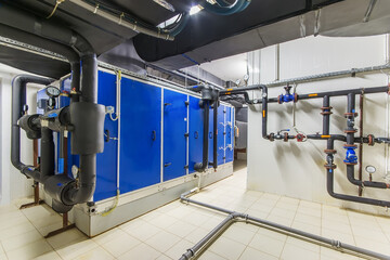 In a spacious industrial facility, blue equipment units are arranged against a wall while complex piping and valves branch out across the floor, showcasing advanced technology and engineering
