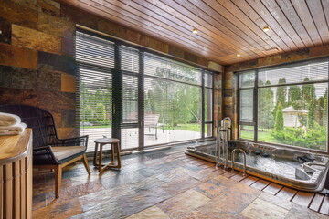 A serene indoor spa featuring a warm hot tub surrounded by natural stone walls, large windows, and a view of a green outdoor space on a peaceful day