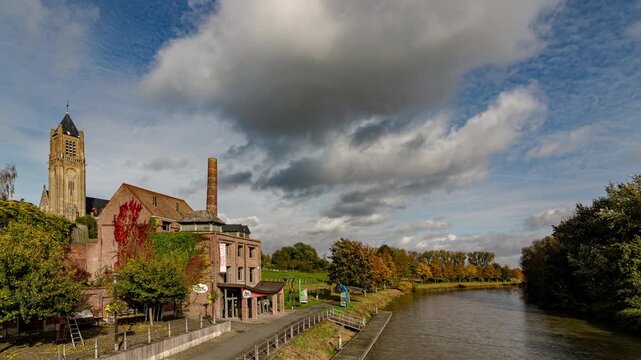Timelapse of a river flowing past an old brick factory chimney and church under a dramatic sky with moving clouds in Warneton village, Belgium