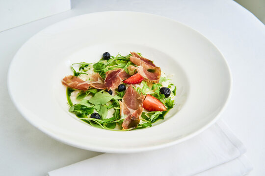 A beautifully plated dish features creamy risotto topped with slices of jamon, vibrant arugula, and fresh strawberries, creating a delightful balance of flavors. Perfect for a gourmet meal