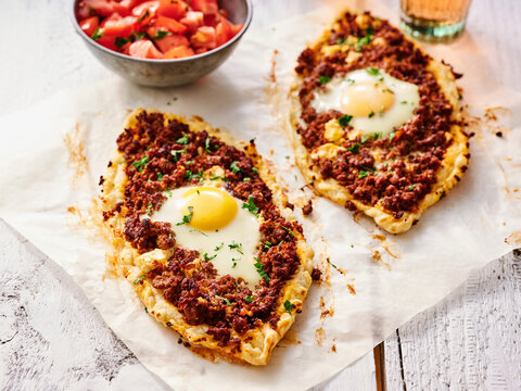 Savory pide topped with ground meat and egg sits on parchment paper alongside a bowl of fresh tomato salad. This dish is enjoyed at a street cafe in Turkey during the busy lunch hour