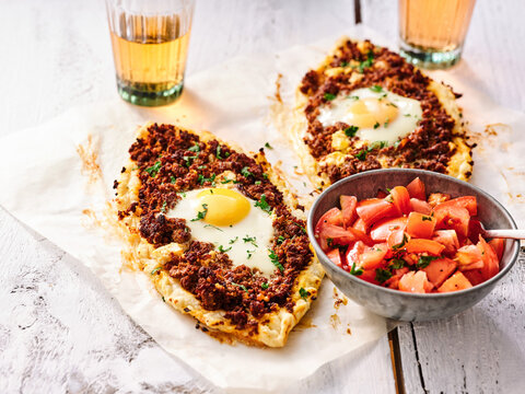 Pide, a Turkish flatbread pizza topped with minced meat and an egg, is served alongside a bowl of fresh tomato salad. Refreshing drinks are also present, enhancing the meal