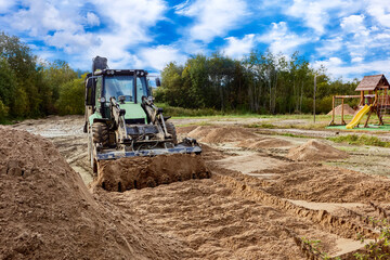 Backhoe loader performs earthmoving work with sand piles visible on site close to playground area.