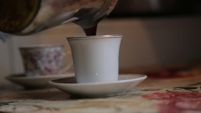 Pouring dark  coffee from  into small, antique porcelain cups. Cozy morning preparation ritual on a floral tablecloth. Delicious hot drink.