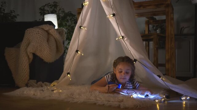 Girl reads with flashlight inside tent and cozy blanket in bedroom, child smiling under fairy light, reading book by flashlight, indoor play with glow, tent and blanket creating snug cozy hideaway