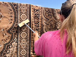  A young woman in a pink T-shirt with tied-back hair is cleaning a hanging carpet outdoors using a brush.