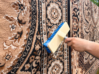 A close-up of a hand holding a brush, scrubbing a hanging carpet outdoors.